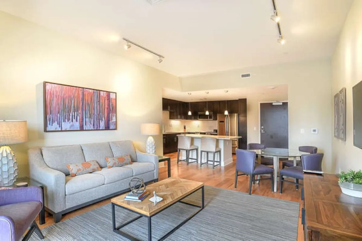 The image showcases a spacious, stylish open-concept living area with a plush sofa, striking local artwork, a sleek kitchen island, and an inviting dining table—perfect for both relaxation and entertaining. The comfortable atmosphere and premium finishes invite social gatherings as well as quiet, cozy moments.