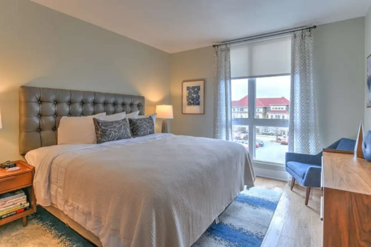 This image features a king bedroom with a plush bed adorned with decorative pillows, a cozy armchair nestled by a window, and a soft influx of natural light. The nightstand and minimalistic decor foster a peaceful, welcoming atmosphere ideal for unwinding or enjoying a book while taking in mountain views.