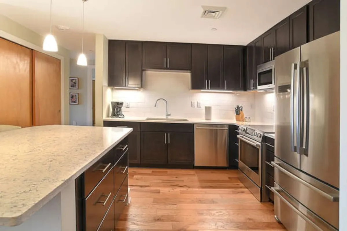 This image captures a different angle of the kitchen, highlighting the large island countertop, stainless steel appliances, dark wood cabinetry, and a well-lit sink area. The atmosphere is inviting and designed for both everyday cooking and hosting memorable gatherings with friends or family.