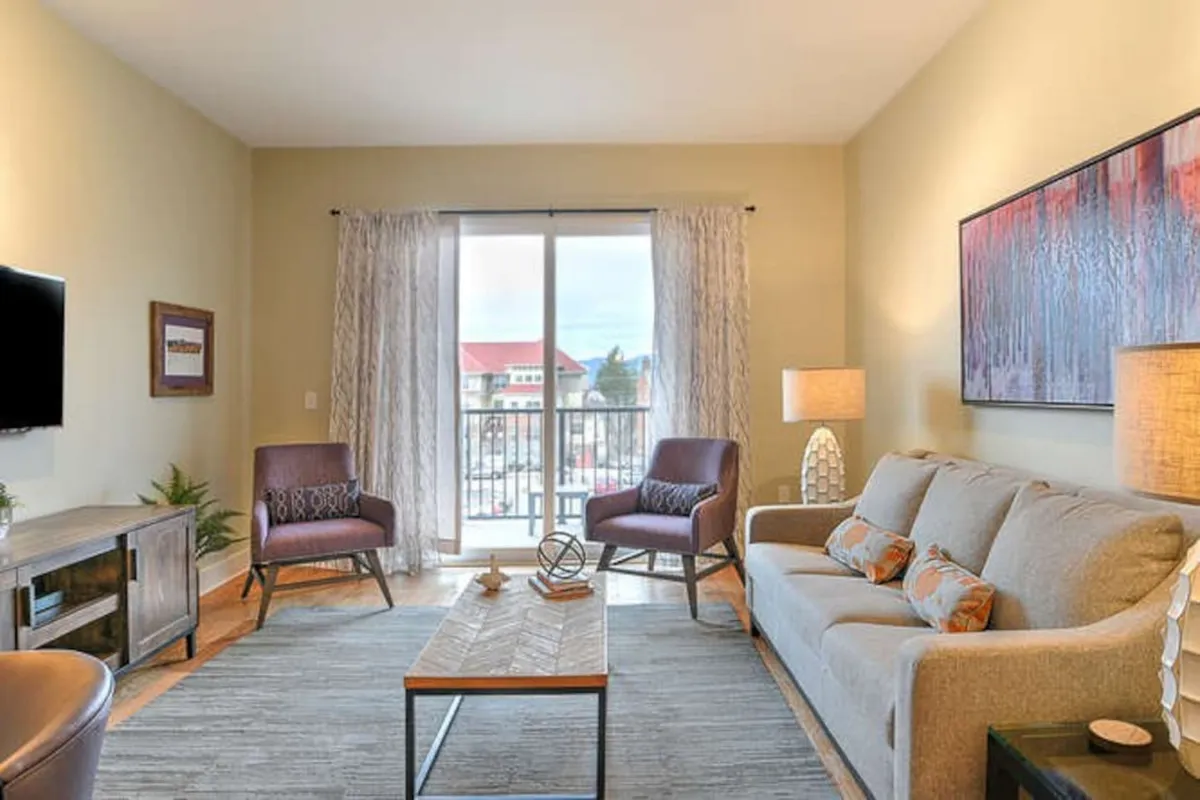 A cozy living area with a plush sofa, coffee table, attractive artwork, and a view through large glass doors that open to a private balcony. The space is styled for both relaxing movie nights and stepping outside to enjoy mountain views or sunset drinks.