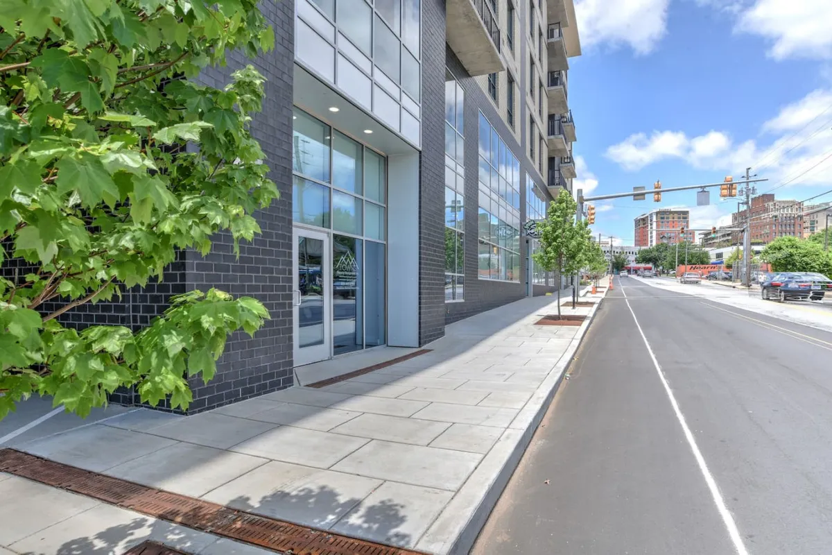 A lively street view featuring a modern building with large glass windows, a tree-lined sidewalk, and people walking, shopping, and dining. The scene has a vibrant urban energy, and hints at the activity and convenience of the downtown area.