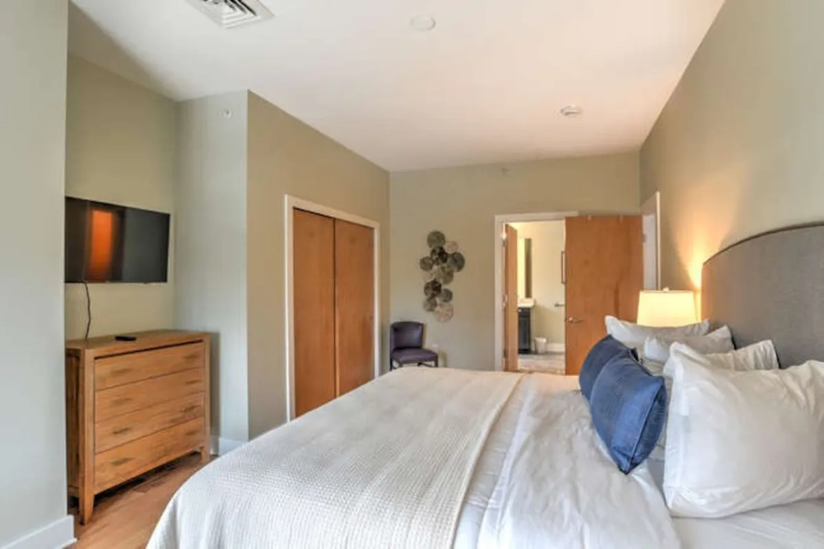 A cozy bedroom featuring a neatly made bed with decorative pillows, a wooden dresser, a wall-mounted TV, an art piece adorning the wall, and a doorway leading to the bathroom. Natural light streams into the room, creating a welcoming atmosphere perfect for relaxing after a busy day of family activities.