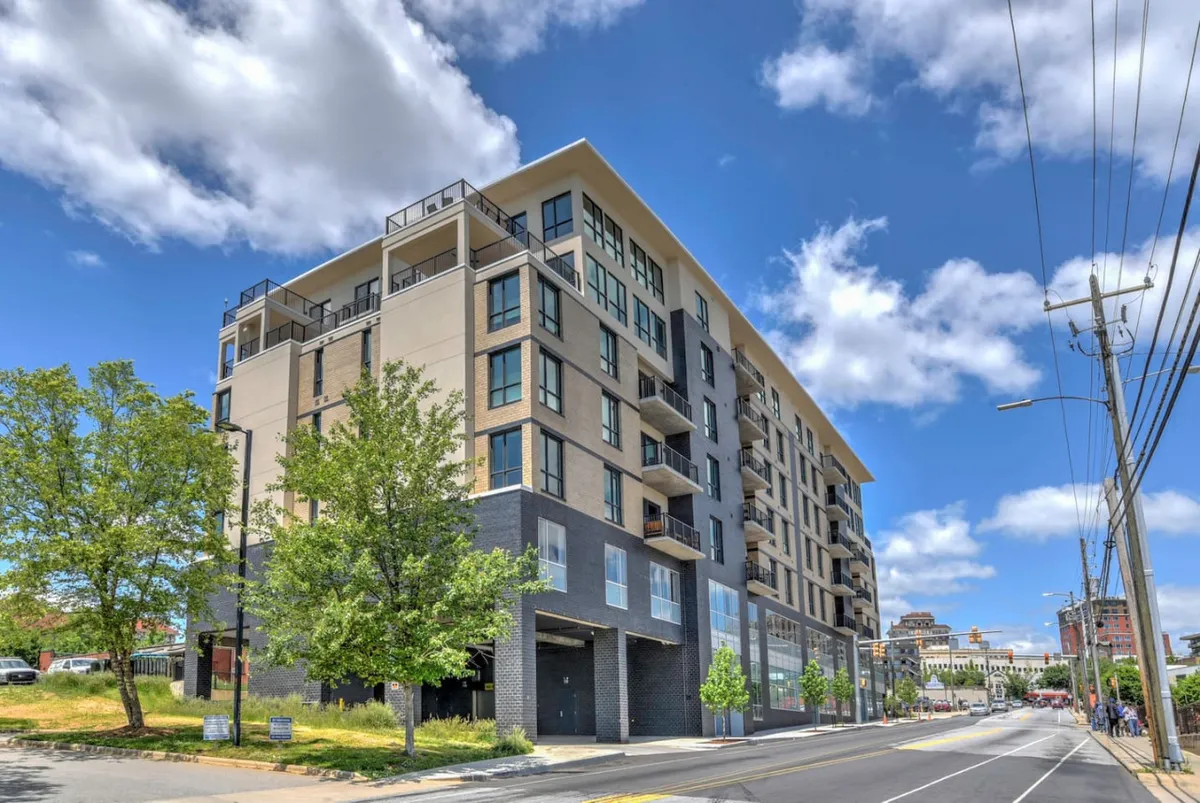 A dynamic street view showcasing modern building architecture surrounded by green trees, with visible sidewalks perfect for walking or biking. The scene is bright and active, highlighting the urban vibrancy and community feel of the neighborhood.