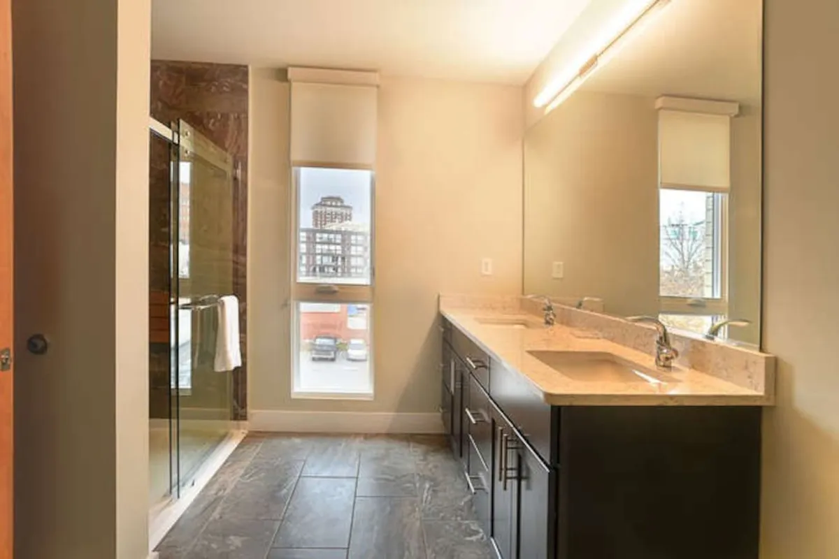 A bright, ADA-accessible bathroom with a double sink vanity, a spacious glass-enclosed walk-in shower with bench seating, and grab bars by the shower for accessibility. Natural light streams through a window, creating an organized and calming atmosphere.
