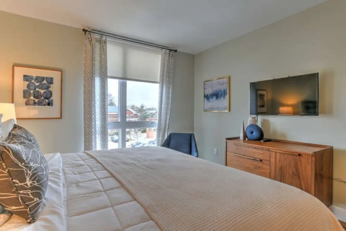 A serene king bedroom setup with a beautifully made bed, sunlight pouring in through modern windows, a wall-mounted cable TV, and a stylish wooden dresser. The setting looks perfect for relaxation, watching TV, or quiet moments in natural light.